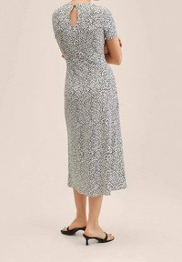 Robe midi à pois noirs et blancs avec manches courtes, détail dos goutte d'eau, et silhouette droite. Portée avec des sandales noires à talons.