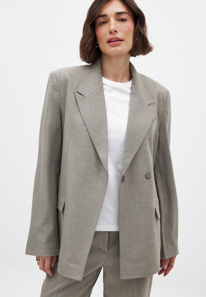 Grau strukturierter, doppelt geknöpfter Blazer mit breiten Revers, seitlichen Taschen und einer lässigen Passform, kombiniert mit einem weißen T-Shirt darunter.