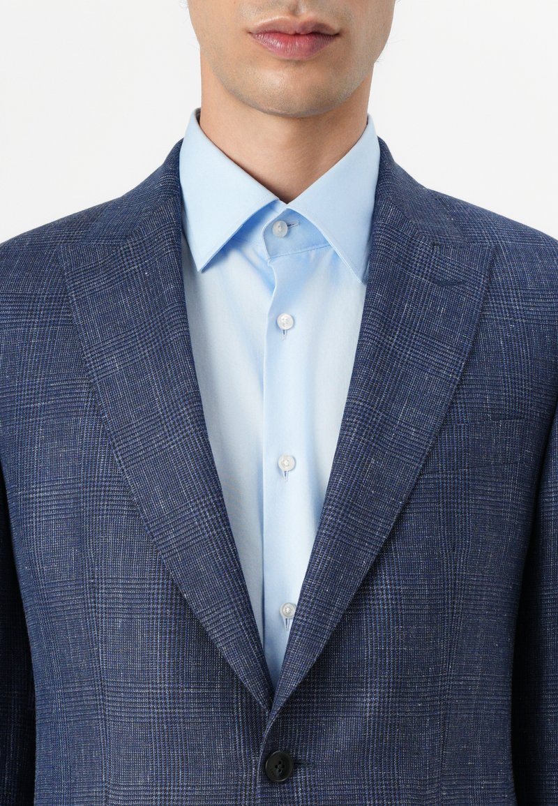 Marineblaues karierter Blazer mit strukturierter Oberfläche, kombiniert mit einem hellblauen Hemd mit Kentkragen und Knöpfen.