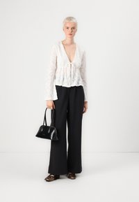Weiße Spitzenbluse mit V-Ausschnitt und Bindedetail, kombiniert mit schwarzen weit geschnittenen Hosen und Mules mit Tierdruck. Hält eine kleine schwarze Handtasche.