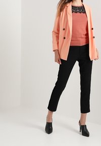 Bledě růžový blazer s dvěma tmavými knoflíky, nošený přes růžový top s krajkovým zdobením, spárovaný s černými kalhotami na míru a černými lodičkami.