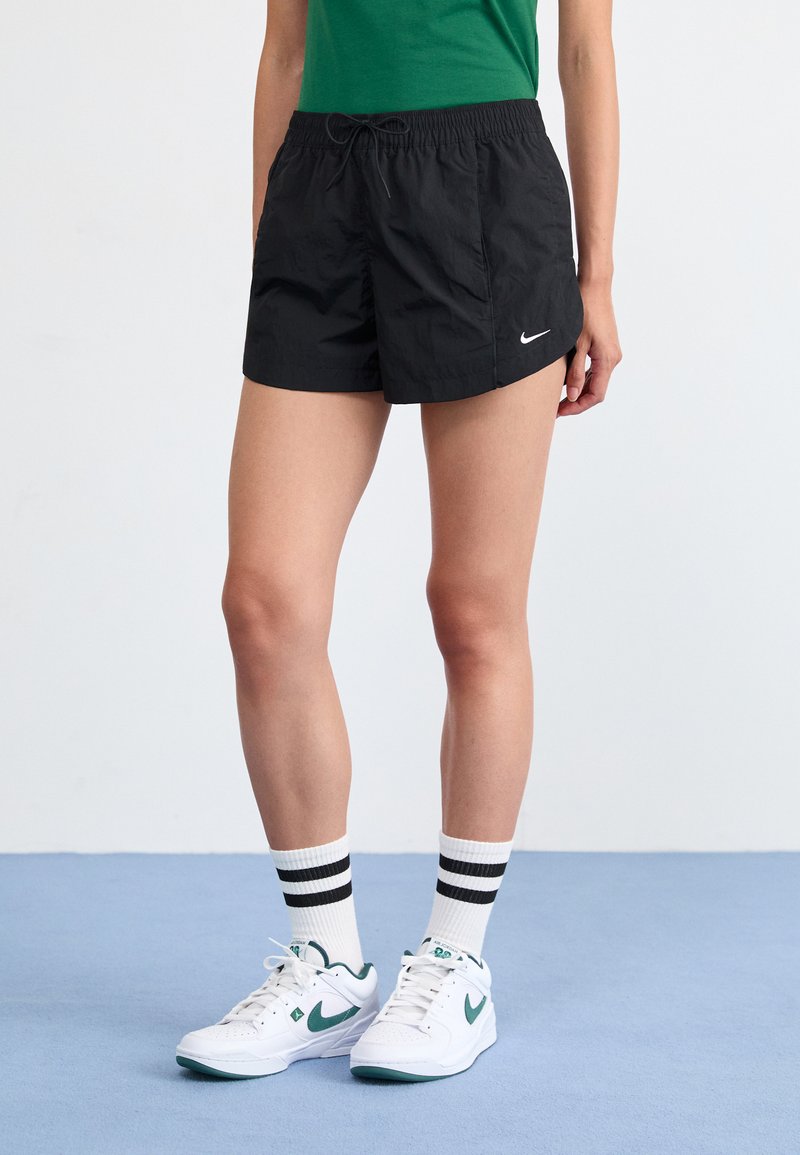 Juodi sportiniai šortai su elastine juostos dalimi, šniūromis ir balta Nike logotipu. Dėvimi su baltais sportiniais bateliais ir juodomis juostelėmis dekoruotomis kojinėmis.