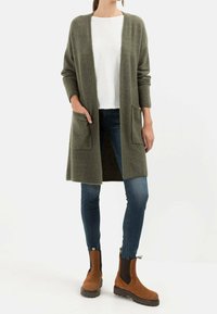 Lang grøn strik cardigan med to forlommer, båret over en hvid skjorte og mørkeblå jeans, parret med brune ankelstøvler.