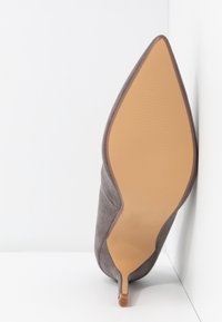 Botte à talon aiguille en daim gris à bout pointu reposant sur sa semelle contre des murs blancs d'angle.