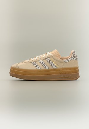 Beige ruskindssneakers med tyk brun sål, leopardmønster på hælen og striber, hvide snørebånd og gyldent "Gazelle" tekst på siden.