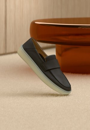 Mocassino nero in pelle con suola bianca spessa, esposto su un tappeto beige accanto a un tavolo rotondo di colore ambra.