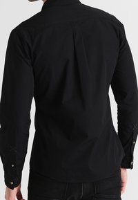 Zwart shirt met lange mouwen, een nauwsluitend ontwerp, een gladde textuur en manchetten met knopen. Heeft een rugbies en subtiele stikseldetails.