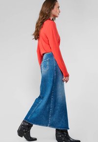 Haut rouge à manches longues associé à une jupe en denim bleue taille haute et longueur cheville. La jupe présente une légère évasement et un détail de poche arrière. Bottines à talons noirs.