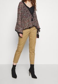 Černý květinový zavinovací top s volánovými rukávy, kombinovaný s světlými úzkými kalhotami a černými špičatými kotníkovými botami. Detaily strukturovaného květinového vzoru.