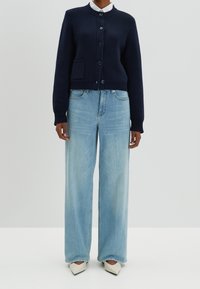 Granatowy sweter z dzianiny zapinany na sześć guzików z kieszonką, w połączeniu z szerokimi jasnoniebieskimi spodniami i spiczastymi białymi butami. Prosty, wygodny krój.