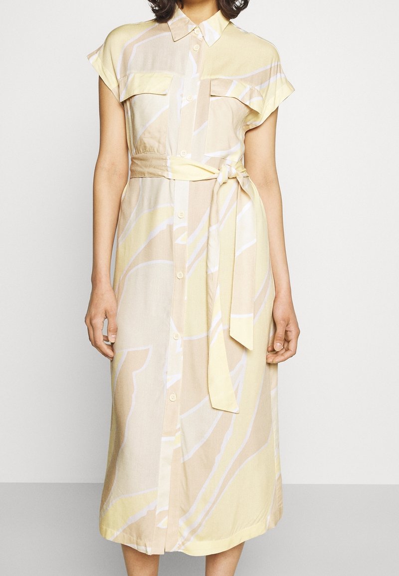 Femme portant une robe-chemise midi jaune pâle et beige avec manches courtes, boutonnière devant, poches poitrine et ceinture nouée à la taille.