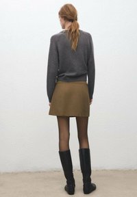 Pull en maille gris, mini-jupe vert olive, collants opaques noirs et bottes noires mi-mollet, vus de dos sur un fond clair.