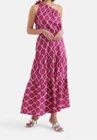 Sukienka maxi w różowym materiale z jednoskrzydłowym krojem. Posiada warstwową spódnicę z falbanami oraz geometryczny wzór w kontrastowych kolorach. Srebrne sandały.