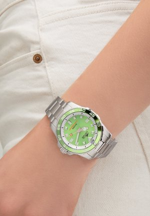 Orologio in metallo argento con quadrante verde, lunetta con accenti neri e indicatori delle ore bianchi. Bracciale in acciaio inox. Visibile su un braccio di colore chiaro.