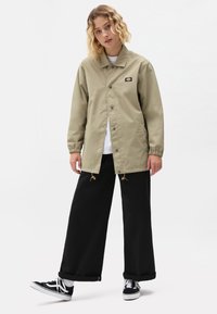 Kaki lett jakke med trykknapper, elastiske mansjetter og snordetaljer, parret med sorte vide bukser og svarte sneakers.