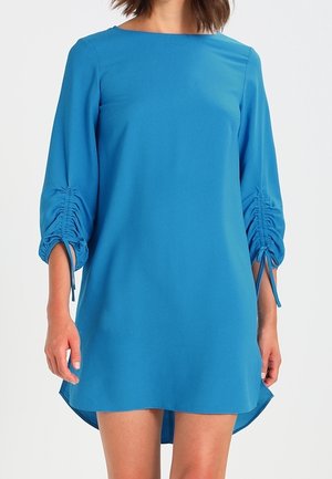 Vrouw draagt een blauw knielang jurk met gerimpelde driekwartmouwen die met touwtjes zijn vastgebonden, staand tegen een effen lichte achtergrond.