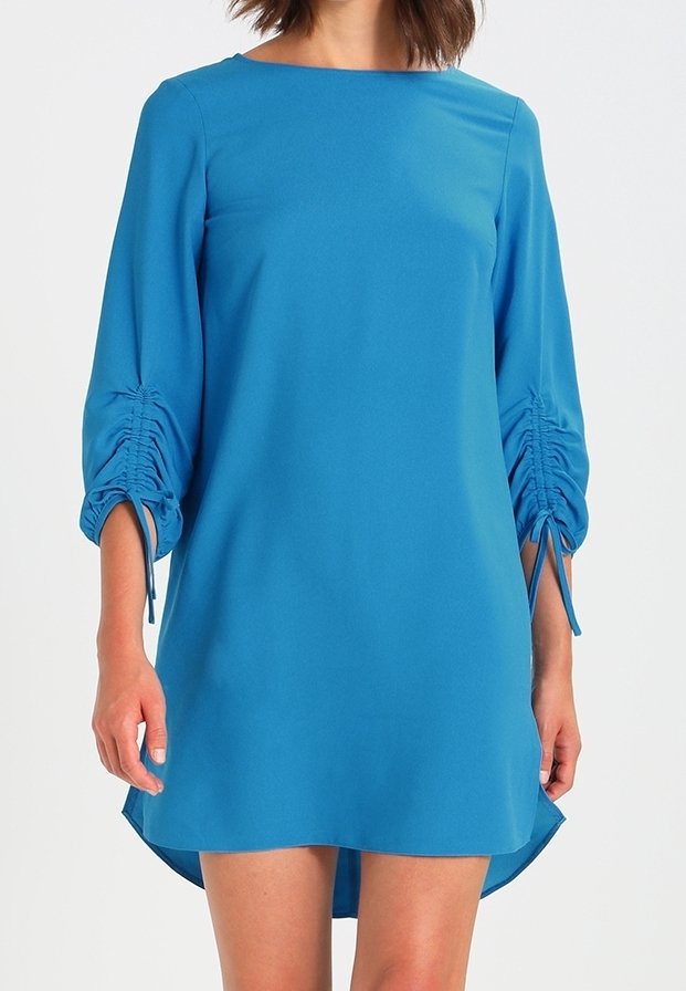 Femme portant une robe bleue d'une longueur au genou avec des manches 3/4 froncées attachées par des cordons, debout devant un fond clair uni.