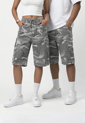 Kaks inimest, kes kannavad halli kaitsevärvi cargo-pükse, valgeid tossusid ja valgeid toppe, seisavad käed taskutes ühevärvilise tausta ees.