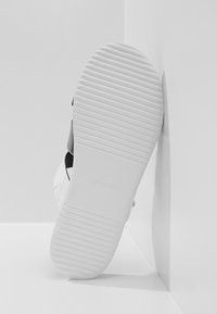 Semelle blanche de sandale Calvin Klein avec des lignes d'adhérence horizontales, appuyée contre un coin sur une surface blanche.