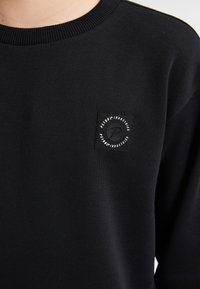 Sudadera negra de cuello redondo con tejido texturizado. Presenta un parche cuadrado negro con el logo en la parte izquierda del pecho. Corte estándar.