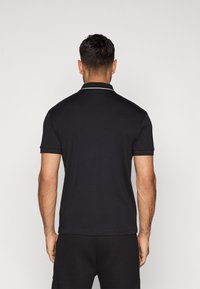 Černé polo tričko s krátkými rukávy, kontrastním bílým límečkem, hladkou texturou a přiléhavým střihem. Nosené s tmavými šortkami.