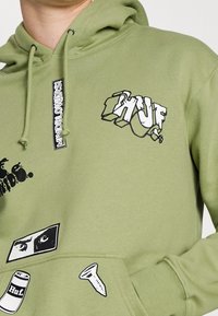 Felpa verde oliva con grafiche frontali: logo "Huf" in testo stilizzato bianco e ulteriori disegni neri, tra cui una vite e un occhio.