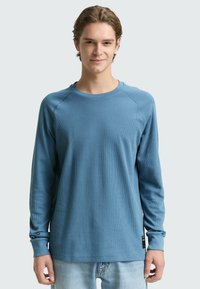 Langärmliges, hellblaues, geripptes Baumwollshirt mit Raglanärmeln und rundem Ausschnitt, mit einem Etikett an der unteren Seite.