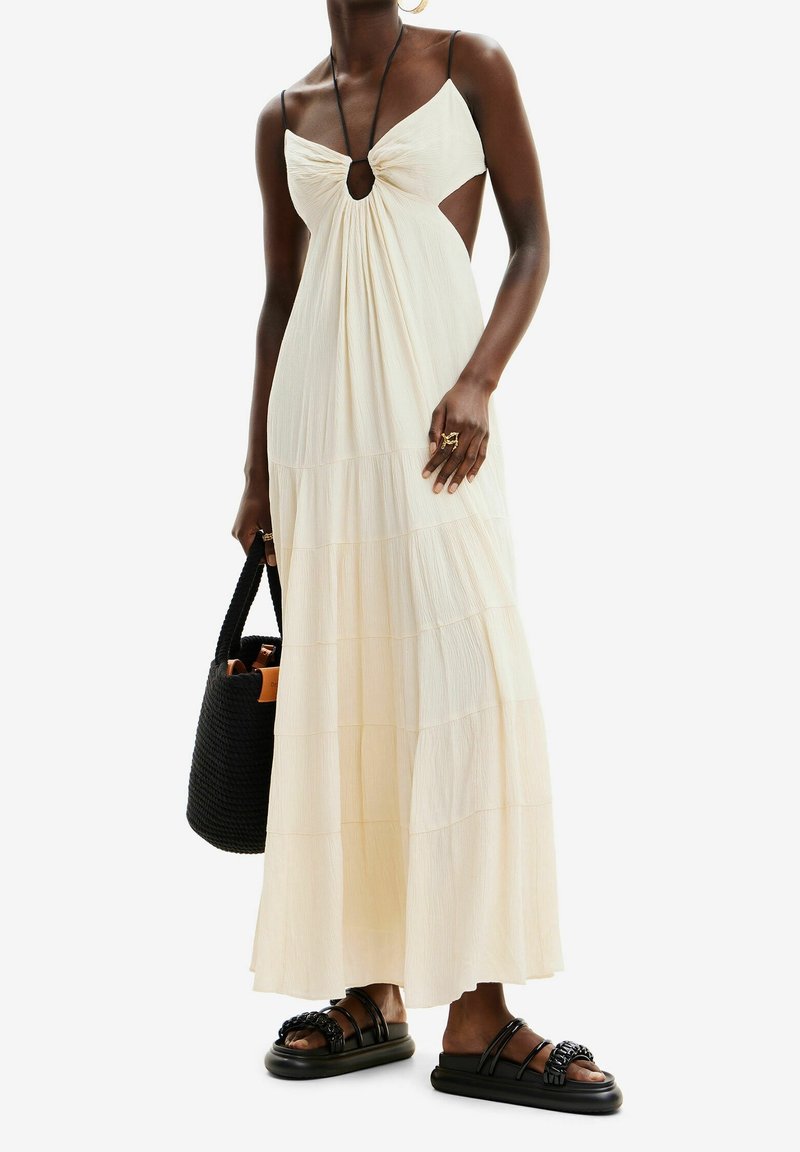 Femme portant une robe maxi crème sans manches avec des découpes, des sandales noires tressées, tenant un sac fourre-tout noir tissé.