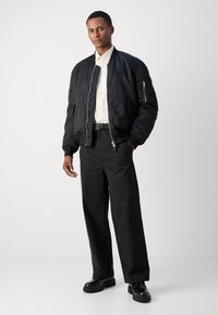 Schwarze Bomberjacke mit gerippten Bündchen, minimaler Markenkennzeichnung, getragen über einem hellen Hemd, kombiniert mit schwarzen Hosen und glänzenden Stiefeln.