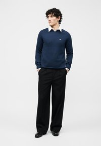 Homme debout, les mains dans les poches, portant un pull bleu marine par-dessus une chemise blanche à col, un pantalon noir à jambes larges et des chaussures noires, sur fond blanc.