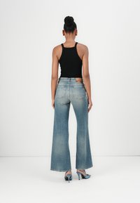 Wide-leg denim jeans in light blue with a faded finish, featuring back pockets and a subtle brand label. Worn with a black tank top and blue heels.