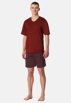 Rotes Baumwoll-T-Shirt mit V-Ausschnitt, kurzen Ärmeln und Brusttasche, kombiniert mit dunkelweinroten, strukturierten Shorts. Model steht barfuß.