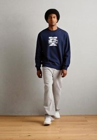 Sudadera marinera con diseño gráfico blanco, combinada con jeans gris claro y zapatillas blancas, llevada por una persona que está de pie frente a un fondo neutro.