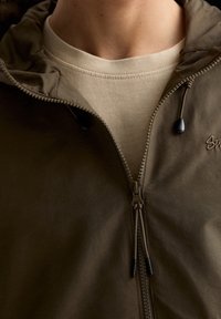 Chaqueta con cremallera en verde oliva con capucha, con acabado texturizado, cremallera frontal y detalles de cordón. Superpuesta sobre una camiseta beige.