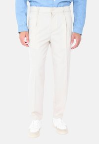 Pantaloni beige con un taglio su misura, caratterizzati da pieghe frontali e una chiusura con zip. Indossati con sneakers bianche e una camicia azzurra chiara.