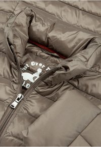 Bruine donsjas met een opstaande kraag, ritssluiting en doorgestikt ontwerp. Heeft een wit logo dat binnenin het kraaggebied is gedrukt.