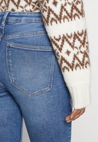 Suéter color crema con patrón geométrico marrón tejido, sobre pantalones vaqueros azules con bolsillo parche y detalle de costuras visibles.