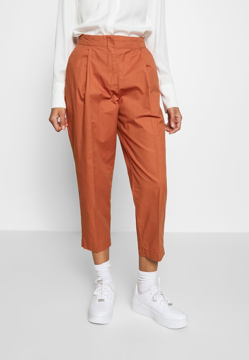 Personne portant un pantalon court de couleur rouille, une chemise blanche à manches longues, des chaussettes blanches et des baskets blanches sur un fond uni.