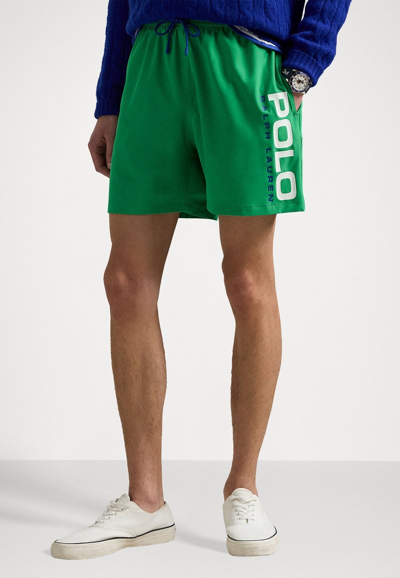 Shorts de bain verts avec une taille élastique, ornés d'un graphique bleu "POLO" sur le côté. Associés à des baskets blanches et un pull bleu.