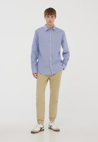 Camicia azzurro chiaro a righe con colletto, abbinata a pantaloni beige a sigaretta e sneakers bianche con dettagli neri.