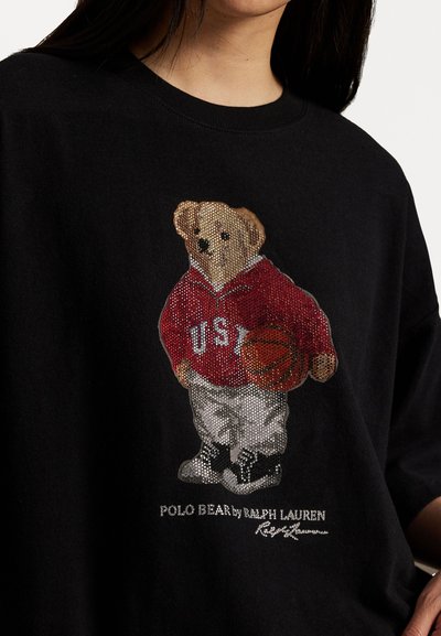 Črna bombažna majica z okrasnim kamnom okrašenim medvedom v rdeči kapuci, z napisom "USA" in košarko, v kombinaciji z belimi hlačami in črnimi športnimi copati.