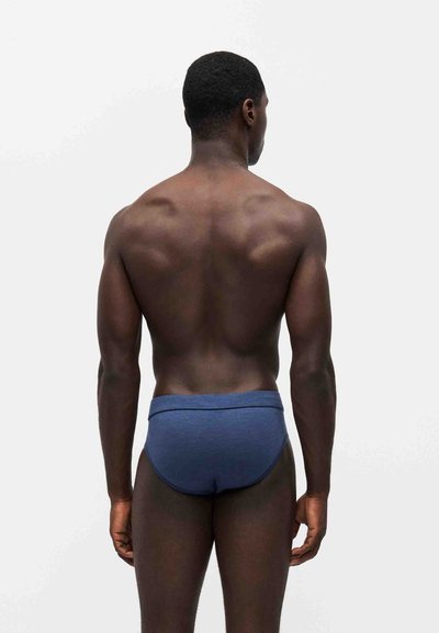 Homme à la peau foncée portant un sous-vêtement bleu, debout dos à la caméra devant un fond clair uni.