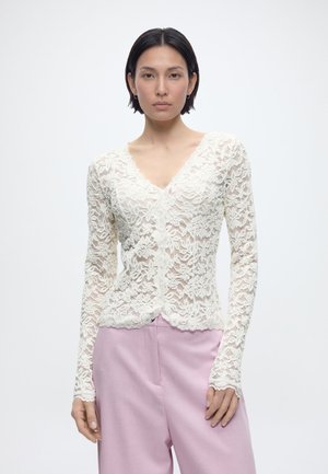 Kvinde med kort sort hår iført en hvid bluse med lange ærmer og blonder samt højttaljede lyserøde bukser mod en ensfarvet baggrund.