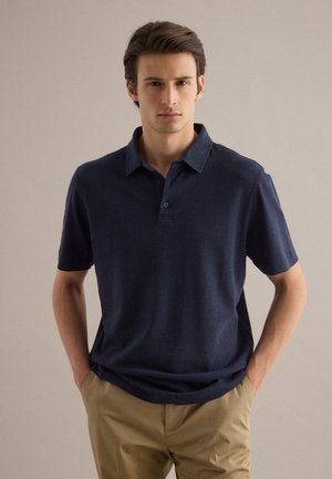 Giovane uomo che indossa una polo a maniche corte blu navy e pantaloni beige, in piedi con le mani nelle tasche davanti a uno sfondo semplice.
