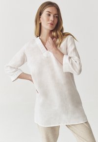 Túnica blanca de lino con escote en V y detalles de encaje intrincados en las mangas. Corte holgado, mangas tres cuartos y dobladillo recto.