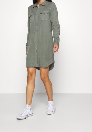 Kobieta w oliwkowozielonej sukience koszulowej z długim rękawem, zapinanej na guziki, z przednimi kieszeniami i białymi tenisówkami za kostkę, stojąca na szarej podłodze.