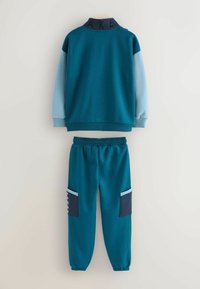 Chándal de dos piezas en teal y azul claro. El pulóver presenta un cuello alto con el texto "AKE". Los pantalones tienen bolsillos laterales y puños de canalé.