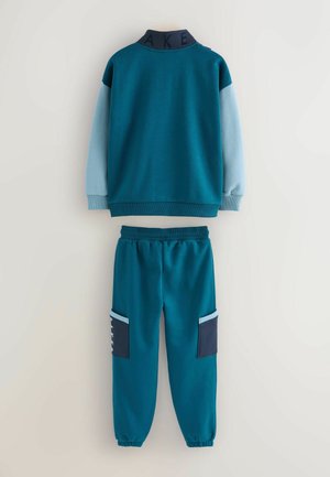 To-delt tracksuit i teal og lyseblå. Pulloveren har en høj krave med "AKE" tekst. Bukserne har sidelommer og ribbede manchetter.