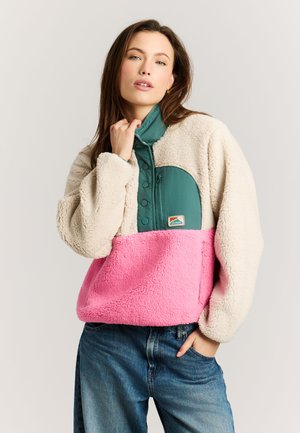 Vrouw draagt een fleecejack met kleurvlakken in beige, groen en roze, staand met een hand aan haar kraag en de andere in haar zak.