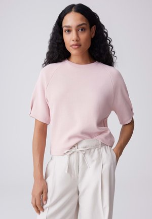 Donna con lunghi capelli ricci che indossa una maglietta rosa chiaro a maniche corte e pantaloni bianchi con coulisse, in piedi davanti a uno sfondo semplice.